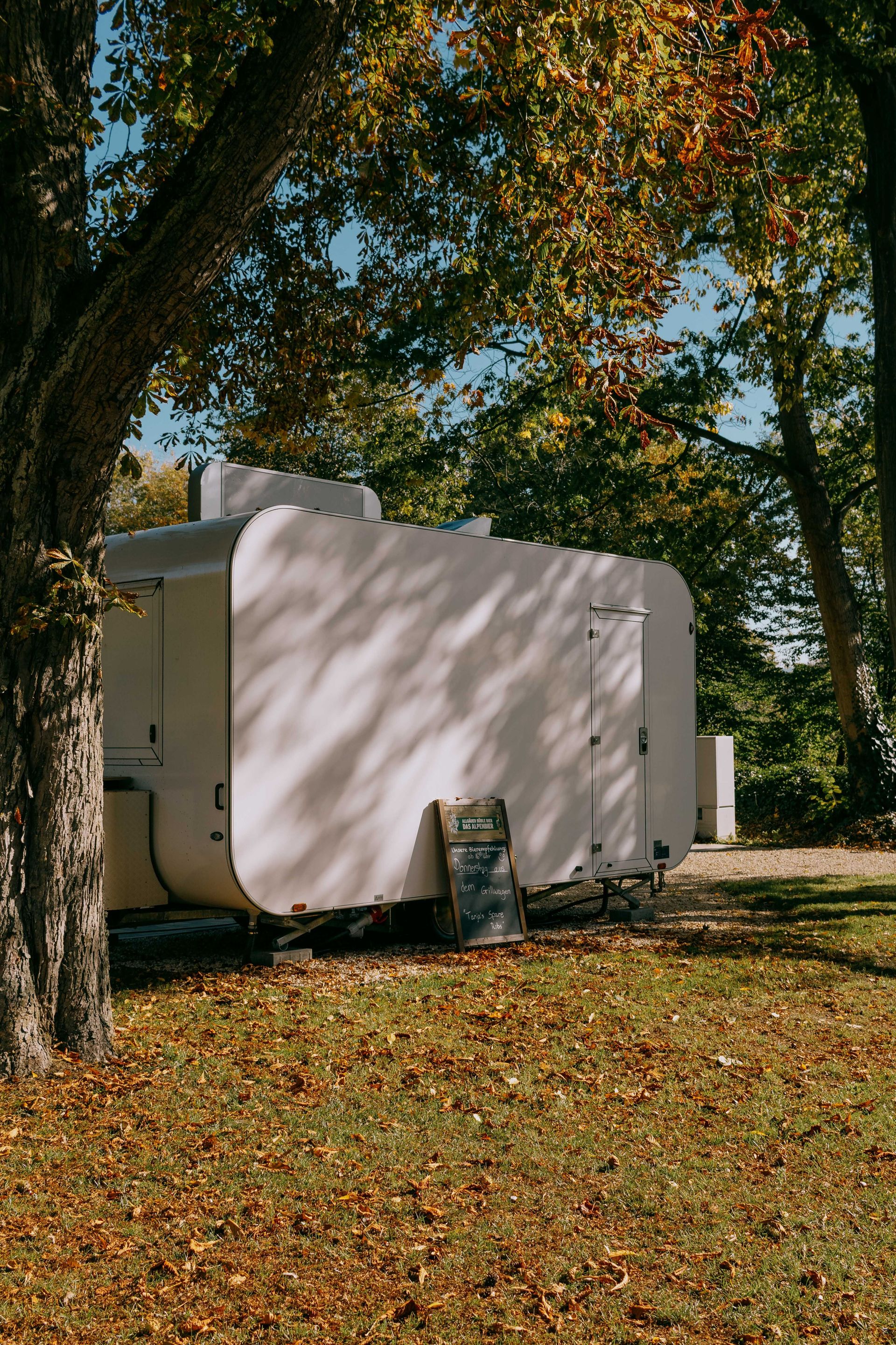 A white caravan parked in a serene forest setting with autumn leaves, offering a cozy outdoor retreat.